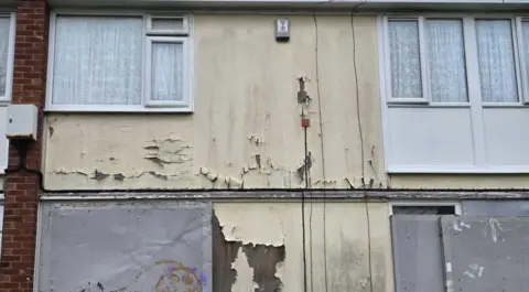 Picture of a boarded up house. Metal sheets cover the windows and paint is coming away from the front of the property.