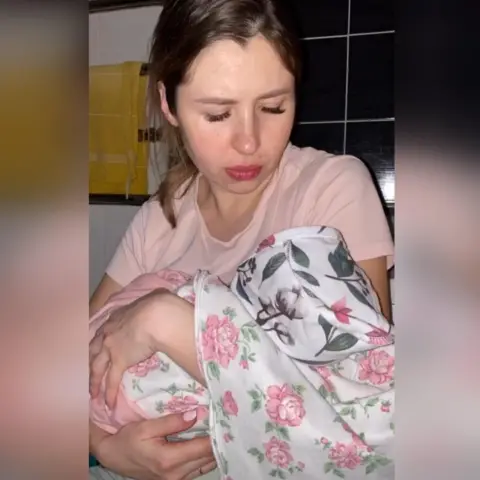 Maryna Korolova Maryna holding her newborn baby. The baby is in a floral onesie and the young woman is in a pink pyjama top. 