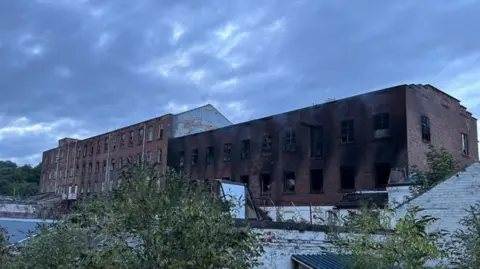 A view of the derelict mill on Andrew Street in Stockport after the blaze. There us black soot around most of the right of the building. A  green bush is in front of a white bricked wall in front of that. The sky is gloomy. 