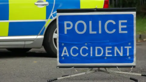 Blue Police accident sign with white writing in front of a police car. Only part of the police car can be seen