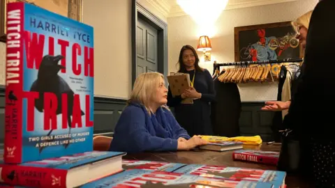 Alex Dunlop/BBC Harriet Tyce sitting down at a table during a book signing event for her latest novel, Witch Trial. The book, coloured blue and red, is in the foreground.