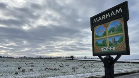 Marham village sign, with paintings of a plane, house, field and church. It stands next to a road and snowy fields, with sheep grazing.