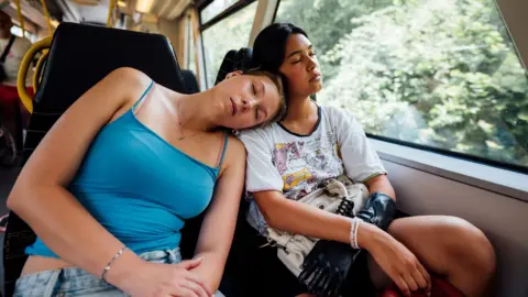 Two young women asleep on a train - one wearing a blue vest top leans on the other's shoulder