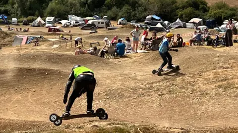 Two mountainboarders race down a hilly track. Spectators can be seen in the background, with campervans and tents behind them. 