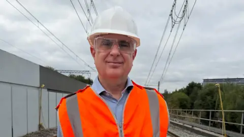 Chris Barnes is standing in front of the tram track where the work is taking place, he's wearing an orange high-vis jacket, a blue shirt underneath and safety goggles with a white helmet.