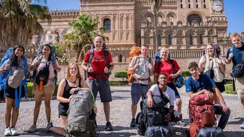 BBC/Studio Lambert Ten contestants stand in front of a historical palace with gardens outside. Each member of the team has a big backpack and wears sports shoes and clothes