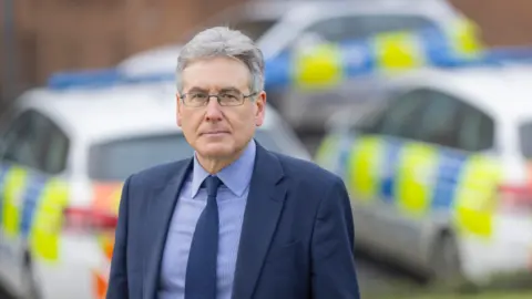 West Midlands Police and Crime Commissioner Mr Foster is standing in front of several police cars which are slightly blurred out. He is wearing a dark suit and navy shirt and tie. He has glasses and grey hair.
