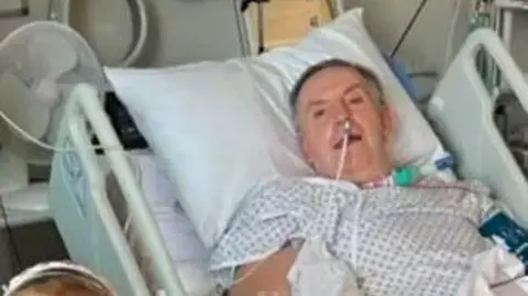 Kevin Pardy in a hospital bed while recovering from Guillain-Barré syndrome. Kevin wears a white hospital gown and has various tubes coming from his face and neck. He is propped up against a white pillow. 