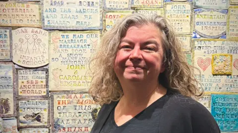 Theresa Heskins, a woman wearing a black top, is standing next to a wall which is decorated with tiles that have various phrases and captions on them. She is looking at the camera and smiling.