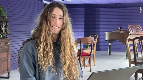 A young man with long curly hair smiles at the camera. He is sitting in a purple room with a piano, tables and chairs that make up the museum's new cafe space. He has a laptop computer in front of him.