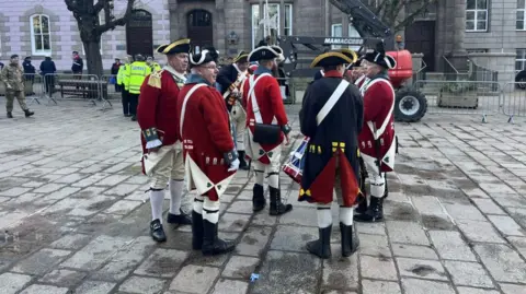 BBC Members of Jersey Militia at the commemoration event last year. The men are dressed in uniforms which are red and white. They are standing on a paved road. Properties are in the background. 