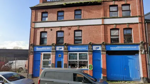 The front of the Shrewsbury Ark building. It is red brick and two storeys, with a blue doors, garage and windows. There is white writing above the doors that says "The Shrewsbury Ark"