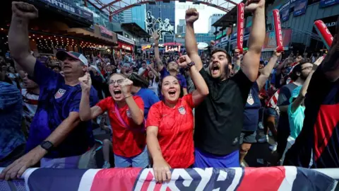 Fans dressed in USA shirts and with USA flags cheer with arms aloft