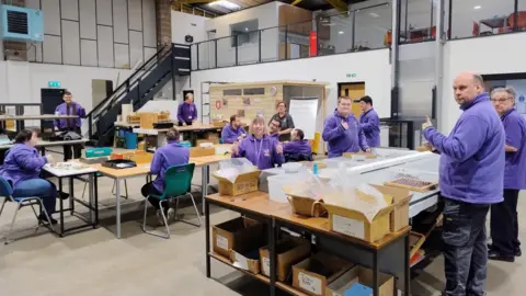 Supported Community Business A group of people wearing purple fleece tops are positioned at various work stations around what appears to be a factory floor. There are boxes on the tables and seats positioned around the tables. There is a set of stairs leading to a balcony area at the read of the scene. There are glass screens and seating areas on the balcony.