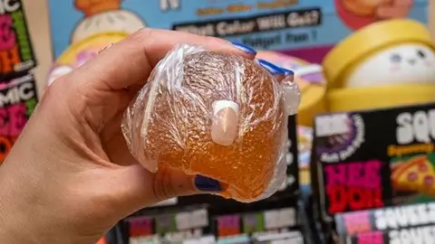 An orange squishy toy is being held, and a false fingernail is protruding from it.