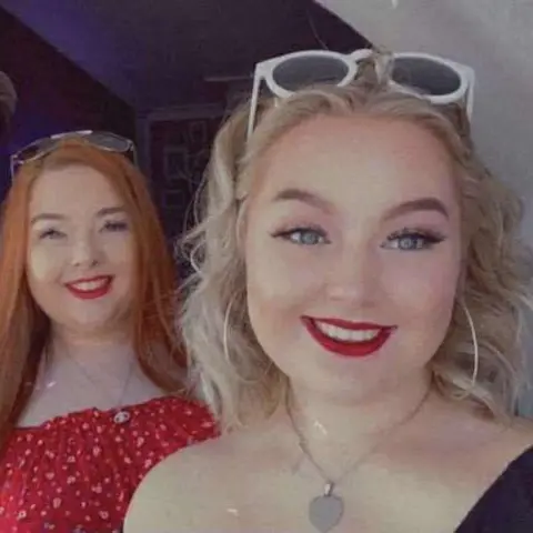 Jessica Thomas Two girls with sunglasses on their heads and red lipstick, smiling at the camera. One has long straight red hair, the other short curly blonde hair. The one with red hair is stood behind the other and has a red top on. They both silver have necklaces with pendants on.