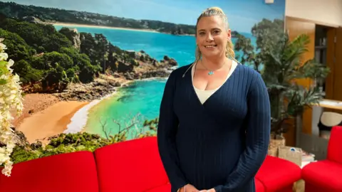 Rachel Boss is standing indoors in front of a scenic coastal backdrop image with turquoise water, rocky outcrops and lush greenery. She has blonde hair and is wearing a dark blue V-neck jumper over a cream top, as well as a silver necklace with a turquoise pendent. Red furniture and white flowers are visible between her and the backdrop.