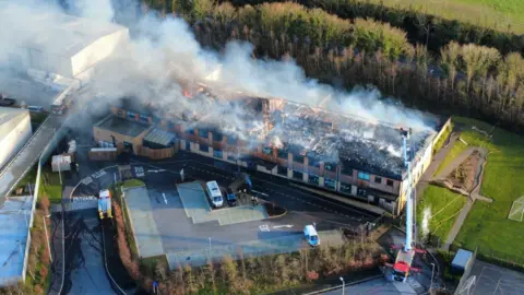A drone image of a school building which is on fire, with a big cloud of smoke coming out of the roof of the building.There is a fire truck and firefighters in the right of the photo.