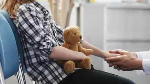 A stock image of a child sitting on a chair.  The image is in profile and shows the child from shoulder height. The child has long blond hair and is wearing a white shirt with a black check, and black trousers.  The child is holding a teddy bear in her right hand and her left hand is being held by a pair of adult sized hands.