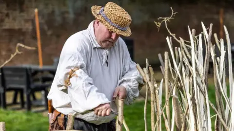 Militias of Anglia A man is wearing a flowing white shirt, and a straw hat. He is slicing through a tin branch of wood with a sharp tool. On his belt appears to be a brown leather tool belt.