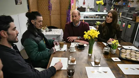 Ben Stansall/PA Wire The Prince and Princess of Wales sit around a wooden table with local residents in Pontypriss. Two men sit on the left with cups of tea. The man wears a green Puffa jacket and has his hair pulled back in a bun; he is talking to Prince William.  Welsh cakes and daffodils sit on the table.