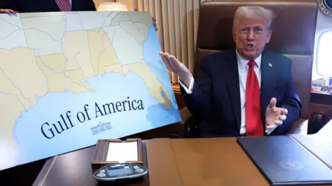 Reuters President Donald Trump sitting at a desk on Air Force One, wearing a navy suit and red tie, gestures towards a placard emblazoned with "Gulf of America"