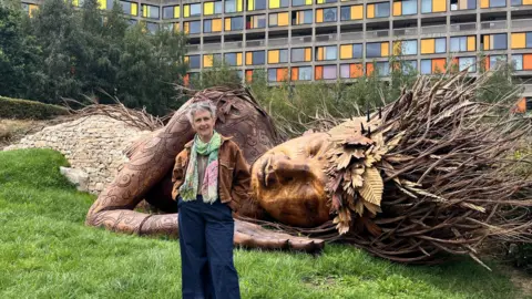 A woman with short grey hair stands in front of a large wooden sculpture of a resting woman. She wears a suede jacket, colourful scarf and dark blue jeans. The wooden sculpture is part of a park installation.
