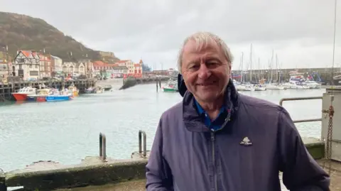 A man with white hair and a blue jacket and polo shirt standing in front of a bay with small boats and buildings in the distance.