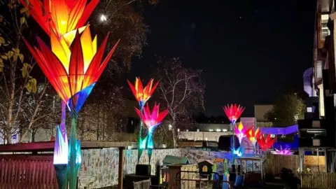 RUH Bath Eight tall flower sculptures which are lit up in orange, red and pink at night. They all have pointed elongated petals pointing up to the sky. Below them is a children's play area at the hospital. 