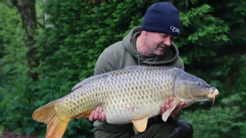 Ross Kaye Ross holding a large fish. He is wearing green trousers, a green hoodie and a blue beanie. He is knelt down with a wooded area behind him.