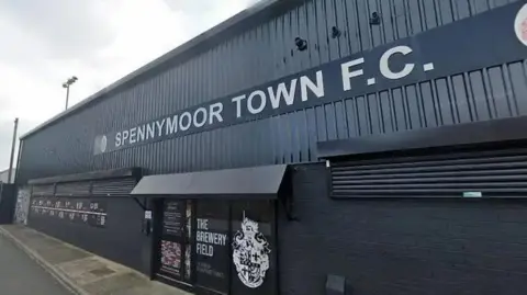 Google A black metal building with a glass door entrance. A sign on the front of the building reads 'Spennymoor Town FC'
