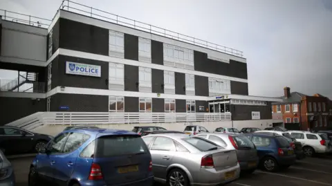 Cowley Police Station, a nondescript box-like building with a "Thames Valley Police" sign, seen beyond a full car park and a white fence lining the walkway to its entrance.