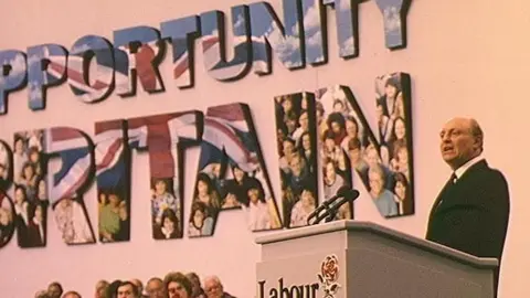 Neil Kinnock on a platform at Labour conference, speaking from a podium with the Labour symbol of a red rose on the front. Behind him on a screen are the words Opportunity Britain, in the colours of the Union Jack