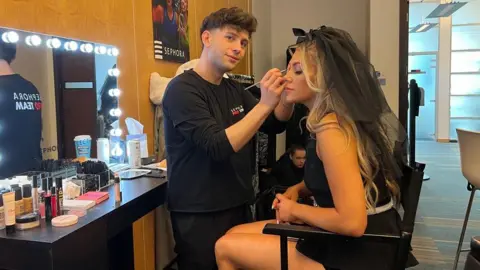 A man has short brown hair and is wearing a black t-shirt. He is standing next to a makeup vanity that is lit up with bright lights. He is putting eye make-up on a woman with long blonde hair. She is wearing a black dress and a black headpiece 