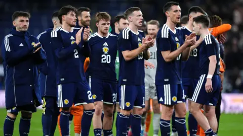Several footballers applauding on the pitch after the end of a match. They are wearing dark blue shirts and shorts