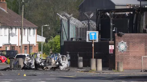 Niall Carson/PA Wire The wreckage of a car bomb outside a police station, with a row of terraced houses and trees in the background. The car has been blown apart and lies on the road beside a set of pavement bollards. The station is protected by a large metal fence, topped with barbed wire, and a red brick wall. 
