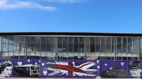 A mural depicting Queen Elizabeth II's likeness in 1952 and 2022, Great Yarmouth's Town Hall, a Union Jack symbol and a a picture of the train that brought her to Great Yarmouth in 1985. The paintings are on a purple background. 
