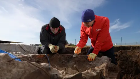 A man in a red top points into a hole. A man in a black jacket is sat next to him, also looking into the hole