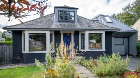 An image of the front of a modernised 1960s bungalow. It is painted navy blue and has a modern side extension in grey materials. Rows of wildflower line the path to the front door which is a rich royal blue.