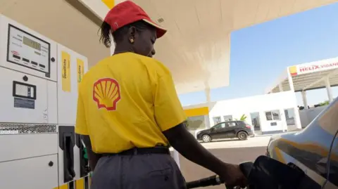 The back of a woman wearing a yellow T-shirt with a Shell logo on the back and a red cap stands by a car as she fills it up with petrol.