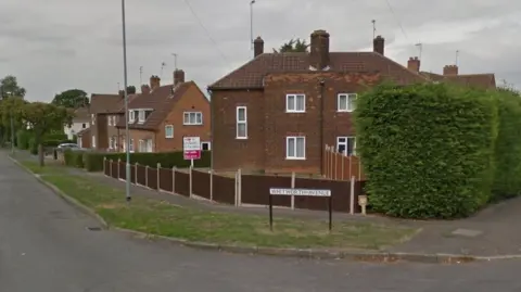 One end of a street of two-storey brick houses with white-framed windows, enclosed by low wooden fences. There is a white street sign saying "Whitworth Avenue" in the centre of the shot. There is a tall hedge to the right and a road with a grass verge sweeps round to the right.