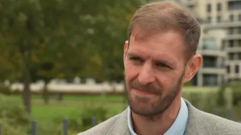 Matt Hughes-Short in a grey suit with a blue shirt. He is standing in a park.