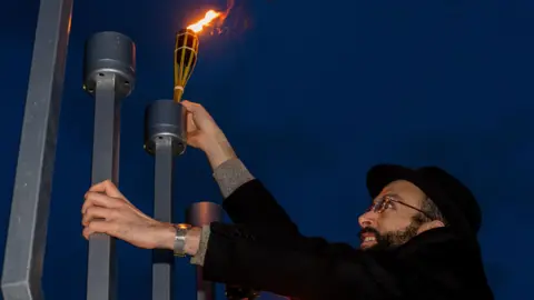Rob Brown, Bristol 24-7 A man is lighting a candelabrum display in the evening. He is wearing glasses, a black hat and jacket. He is lighting up the display using a wooden torch.