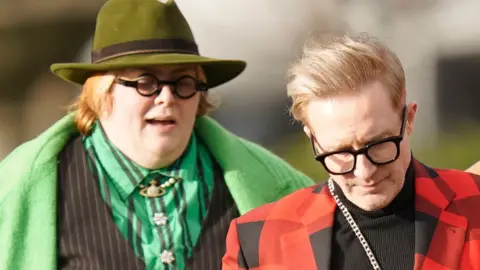 PA Media Comedian Jayde Adams in a green shirt, coat and hat, next to Ian "H" Watkins in black and red checked jacket and black turtleneck