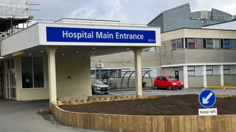 A view of Guernsey's Princess Elizabeth Hospital showing the main entrance, which is signposted with a large blue sign. The building has two storeys and two cars are parked outside.