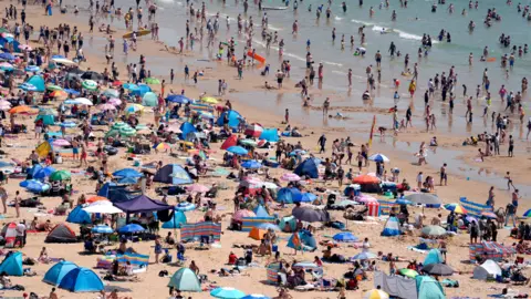 An extremely busy beach on a sunny day. Lots of umbrellas, windbreakers, people sunbathing and people in the sea