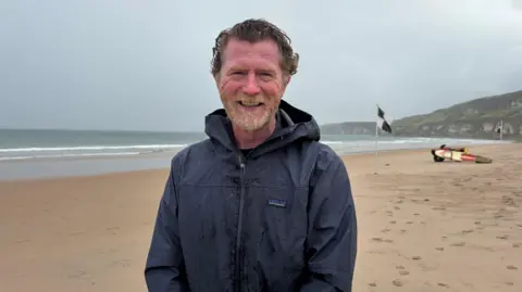 BBC Andrew Todd stands on Whiterocks beach on Portrush. He has wavy brown hair and a ginger/grey beard. He is wearing a dark blue raincoat. It is an overcast day.