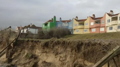 Jamie Niblock/BBC Several homes can be seen at the top of a cliff edge. The homes are all different colours. A crumbling cliff edge can be seen below them.