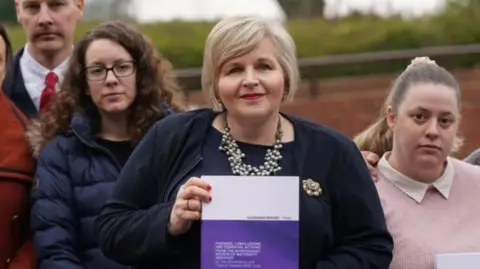 A woman with bobbed blonde hair is wearing a chunky necklace, flower broach, black top and black cardigan. She is standing in front of a gathering of woman and is holding purple and white book in her hands that says report on it. 