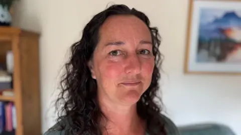 A woman with long dark curly hair wearing a khaki coloured top